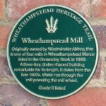 A plaque on a wall for Wheathampstead Heritage trail reading “Wheathamptstead Mill. Originally owned by Westminster Abbey, this is one of four mills in Wheathampstead Manor listed in the Domesday Book in 1086. A three-bay timber framed building remarkable for its length, it dates from the late 1500s. Water ran through the mill powering the mill wheel. Grade two listed.”